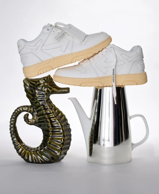 Elevated view of stacked white sneakers, silver teapot, and green seahorse figurine against a white backdrop in a studio setting.