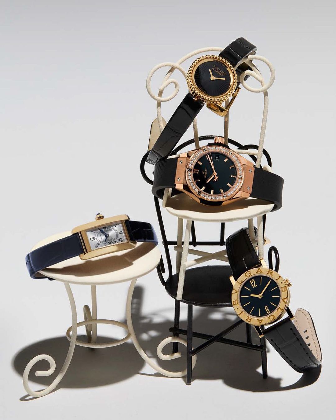 Collection of luxury wristwatches arranged on miniature furniture in a studio setting, showcasing various brands and designs.