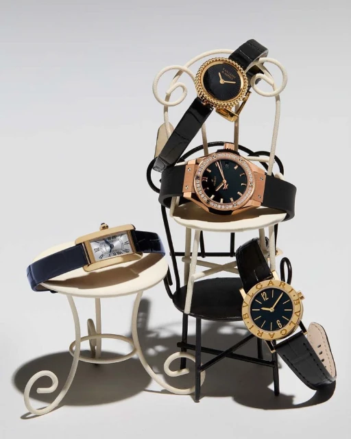 Collection of luxury wristwatches arranged on miniature furniture in a studio setting, showcasing various brands and designs.