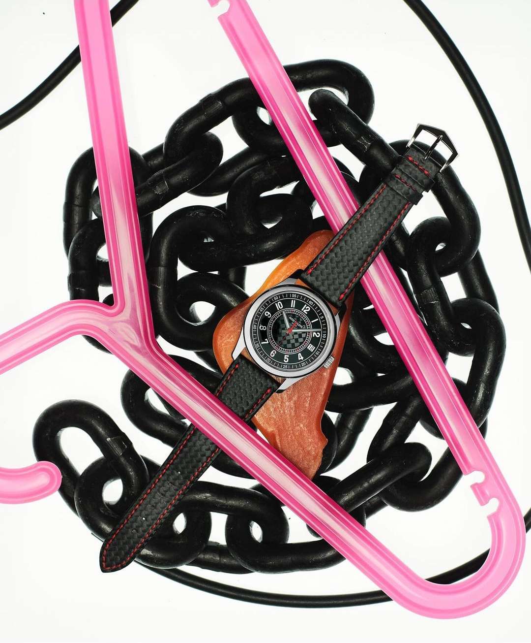 Wristwatch on a brown surface with black chains and pink plastic hangers against a white backdrop in a studio setting.