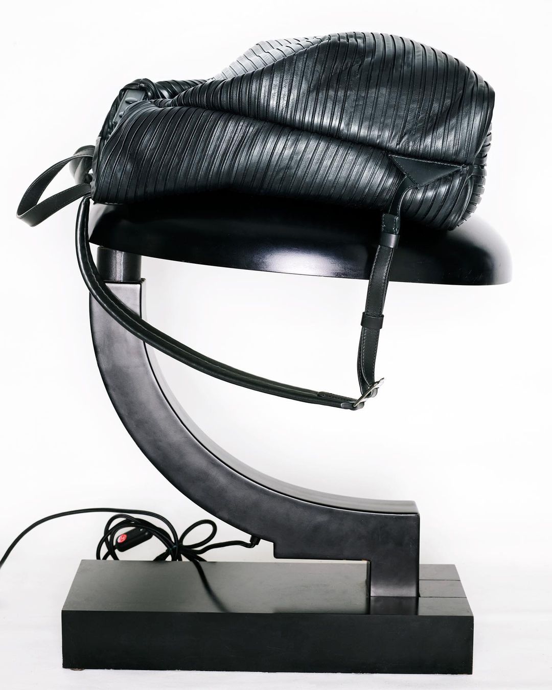 Black leather bucket bag draped over a modern, black art deco style lamp with a white background in a studio product shot.