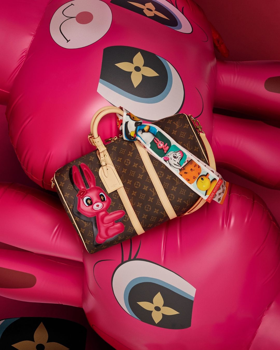Designer handbag with cartoon rabbit charm and patterned scarf against an inflatable pink cartoon bear, in a studio setting.