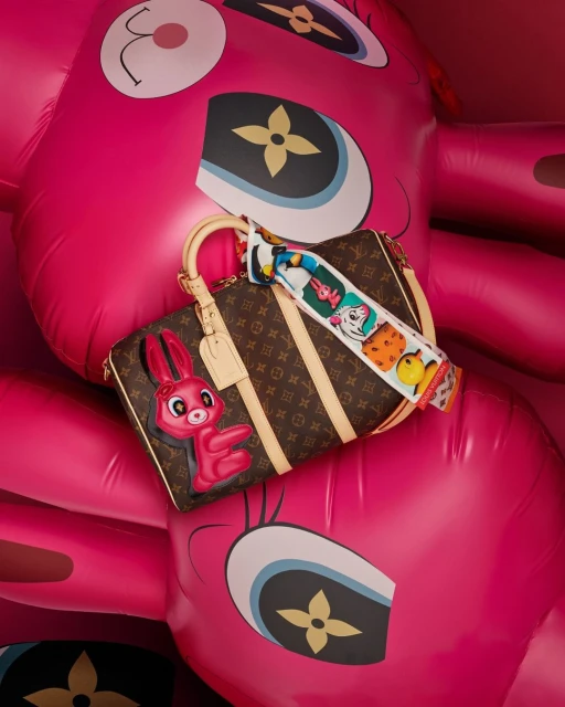 Designer handbag with cartoon rabbit charm and patterned scarf against an inflatable pink cartoon bear, in a studio setting.