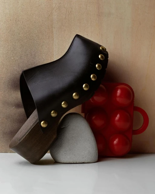 Dark brown leather clog with gold studs, balanced on a gray rock and red plastic object against a beige background in a studio shot.