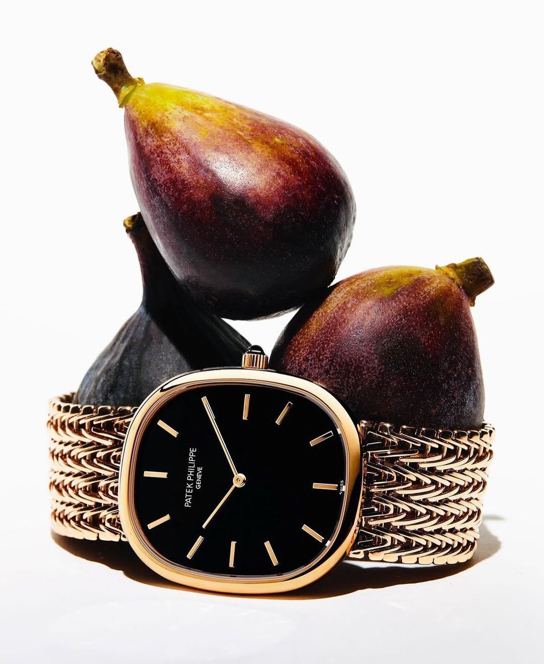 Close-up studio shot of a gold Patek Philippe watch surrounded by dark figs against a bright white background, fashion still life.