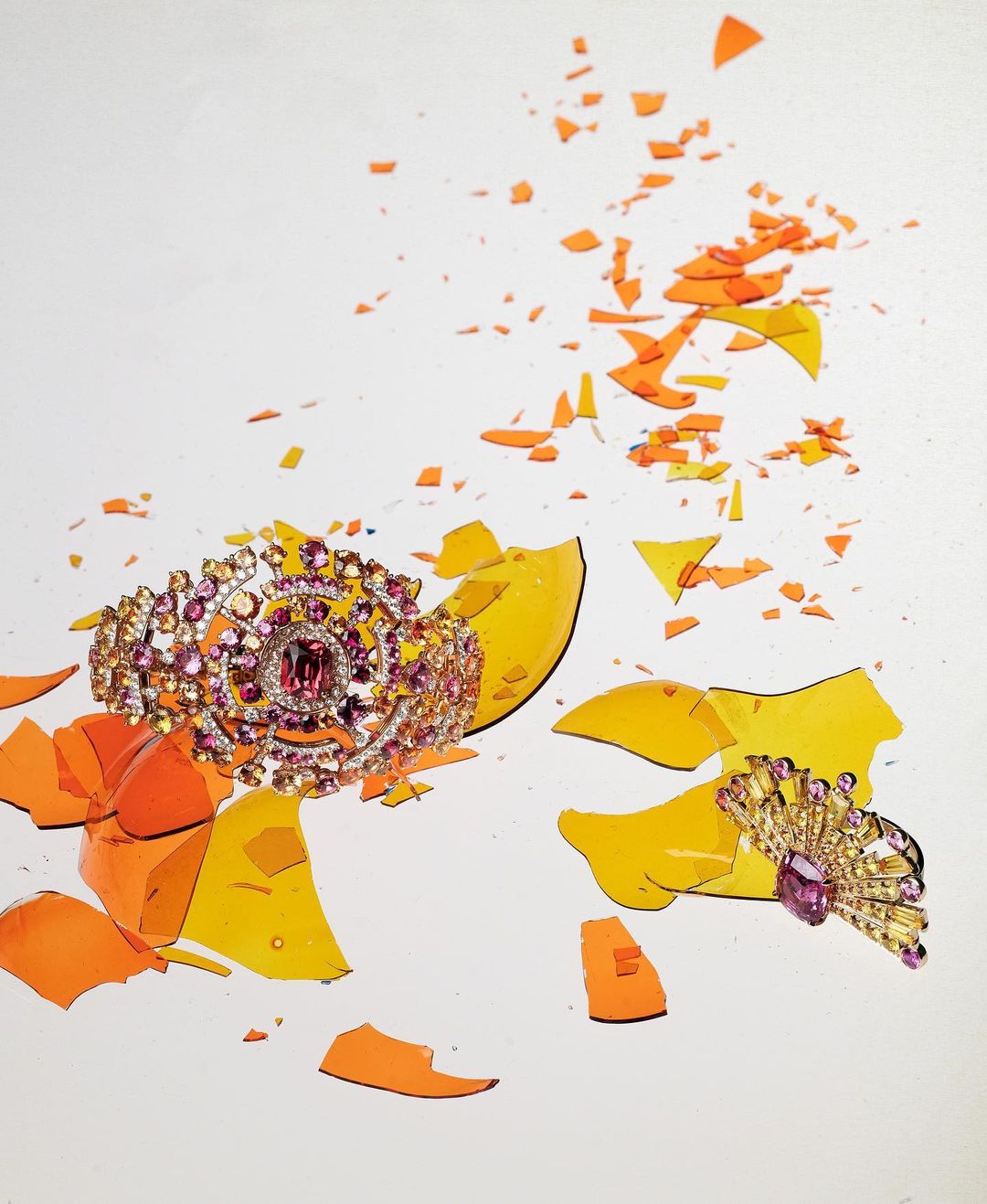 Stunning still life featuring a jeweled crown and brooch surrounded by shattered orange and yellow glass shards against a white backdrop.