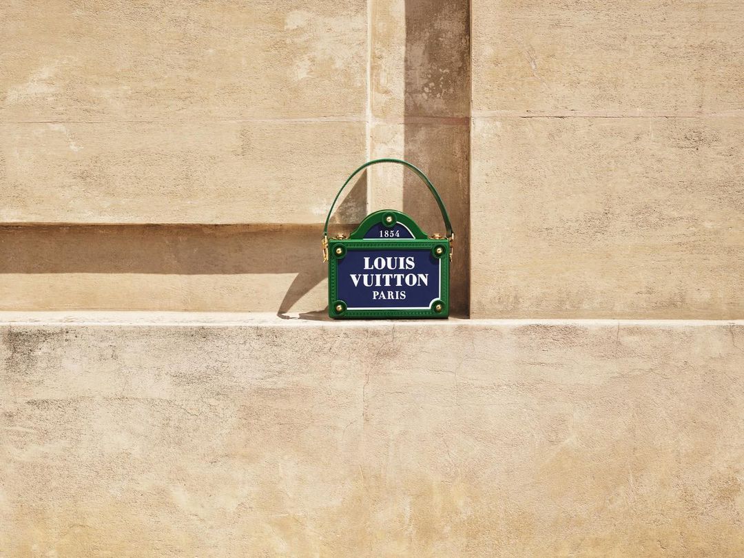 Louis Vuitton purse resembling a street sign, with blue, green, and gold tones against a textured wall. Fashion accessory.