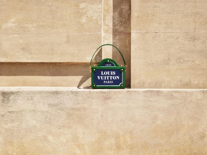 Louis Vuitton purse resembling a street sign, with blue, green, and gold tones against a textured wall. Fashion accessory.