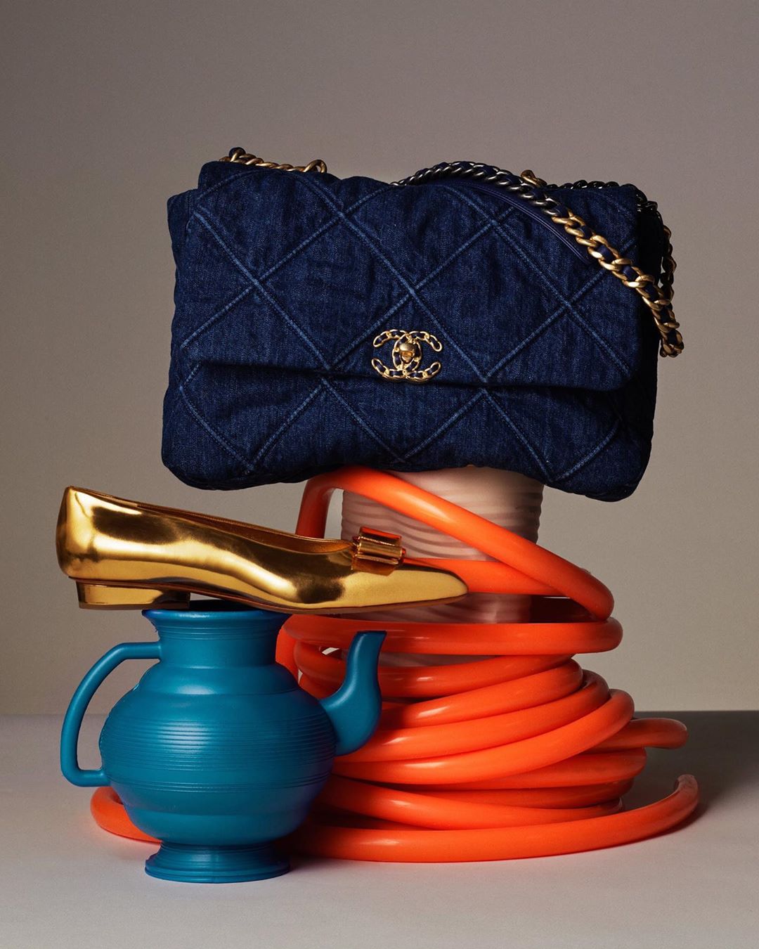 Denim Chanel bag on a still life with a gold shoe, orange tubing, and a blue teapot in a studio setting for a fashion shot.