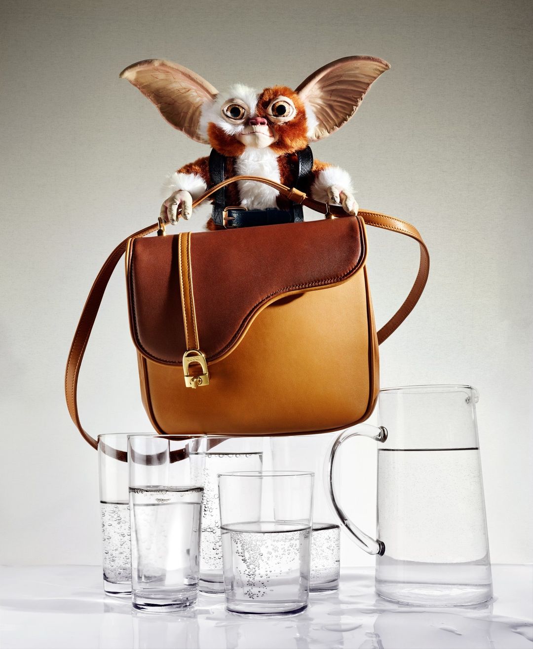 Whimsical studio shot of a brown leather satchel bag carried by a Mogwai-like puppet atop several glasses and a water pitcher, against a neutral backdrop.