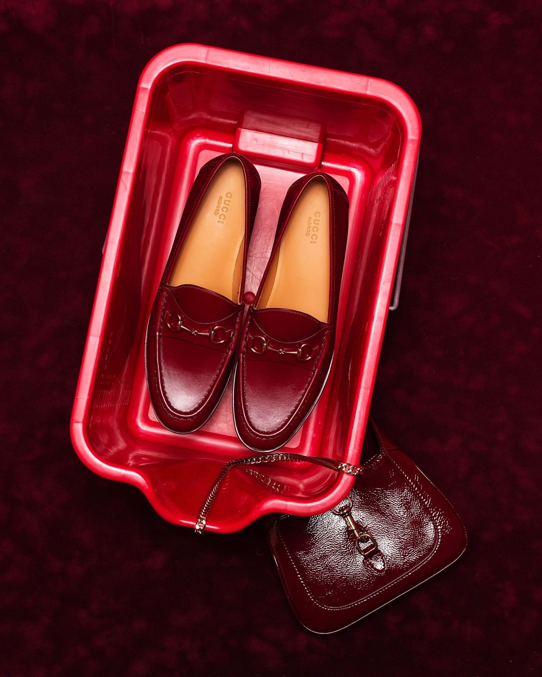 High-angle shot of a pair of Gucci loafers inside a red plastic tub, with a matching purse on a burgundy velvet surface.