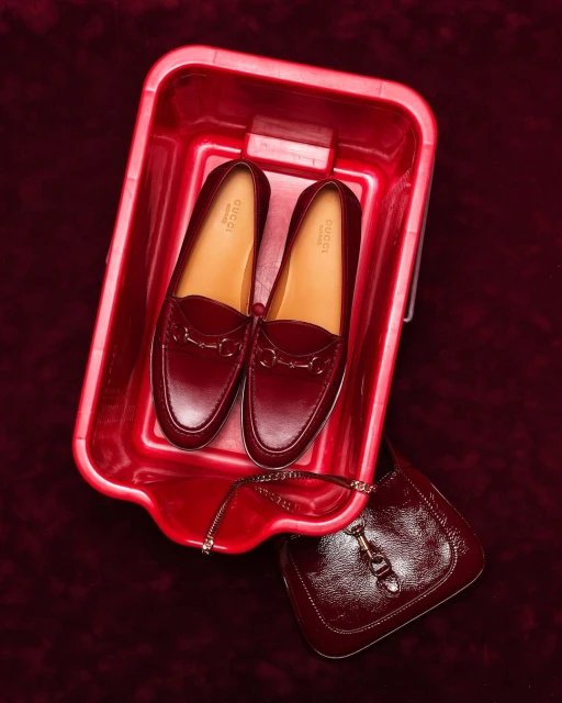 High-angle shot of a pair of Gucci loafers inside a red plastic tub, with a matching purse on a burgundy velvet surface.