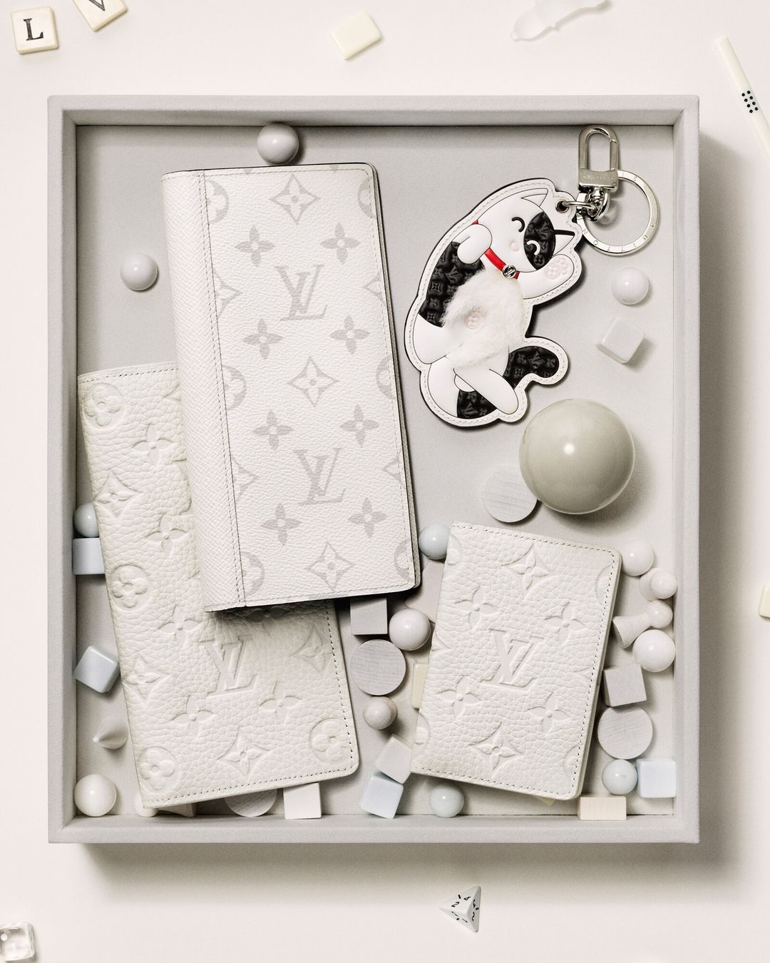 A top-down shot displays white leather wallets, a playful dog keychain, and decorative spheres and cubes on a light grey tray.