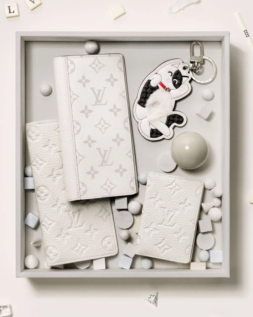A top-down shot displays white leather wallets, a playful dog keychain, and decorative spheres and cubes on a light grey tray.