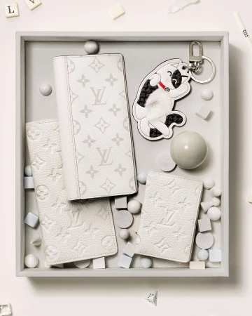 A top-down shot displays white leather wallets, a playful dog keychain, and decorative spheres and cubes on a light grey tray.