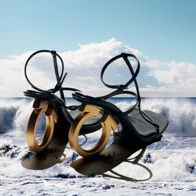Black strappy sandals with gold hardware displayed against a backdrop of ocean waves and blue sky for a luxury fashion aesthetic.
