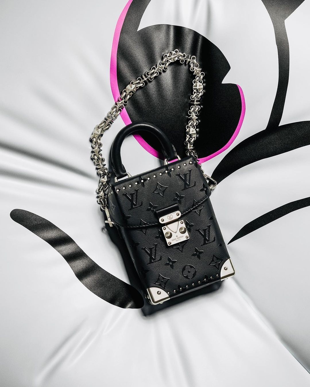 Black Louis Vuitton box bag with a silver chain strap and silver hardware, resting on a white surface with black and pink accents.