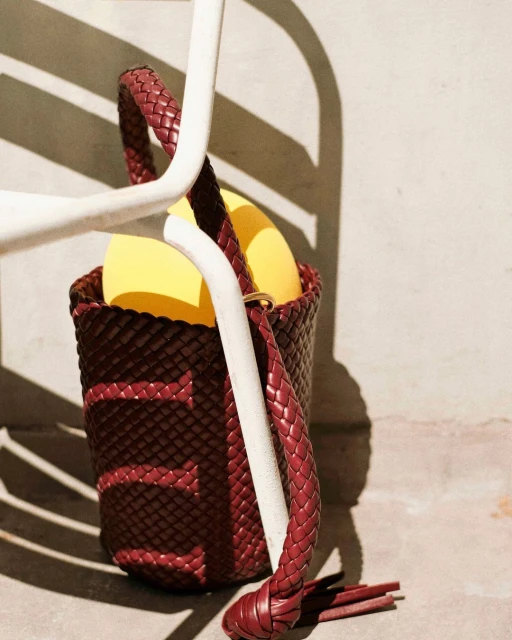 Burgundy woven handbag with yellow liner and braided strap leaning against a white chair with shadows on a beige background.