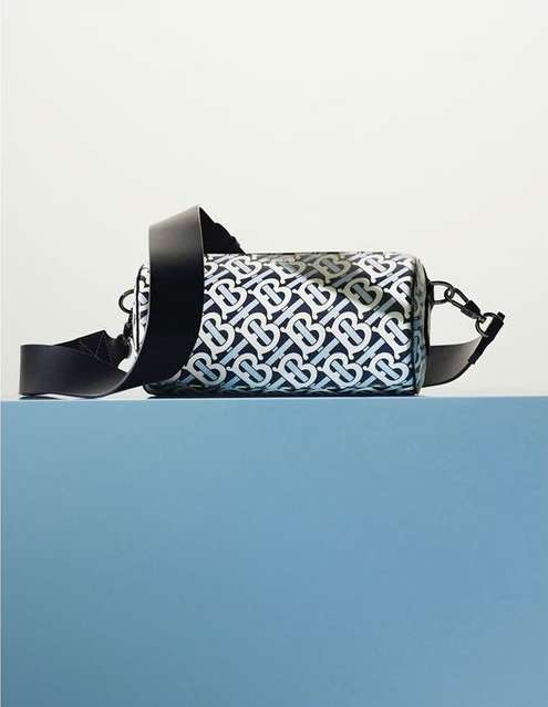 Monogram barrel bag with a thick black shoulder strap on a blue platform against a neutral backdrop in a product studio shot.