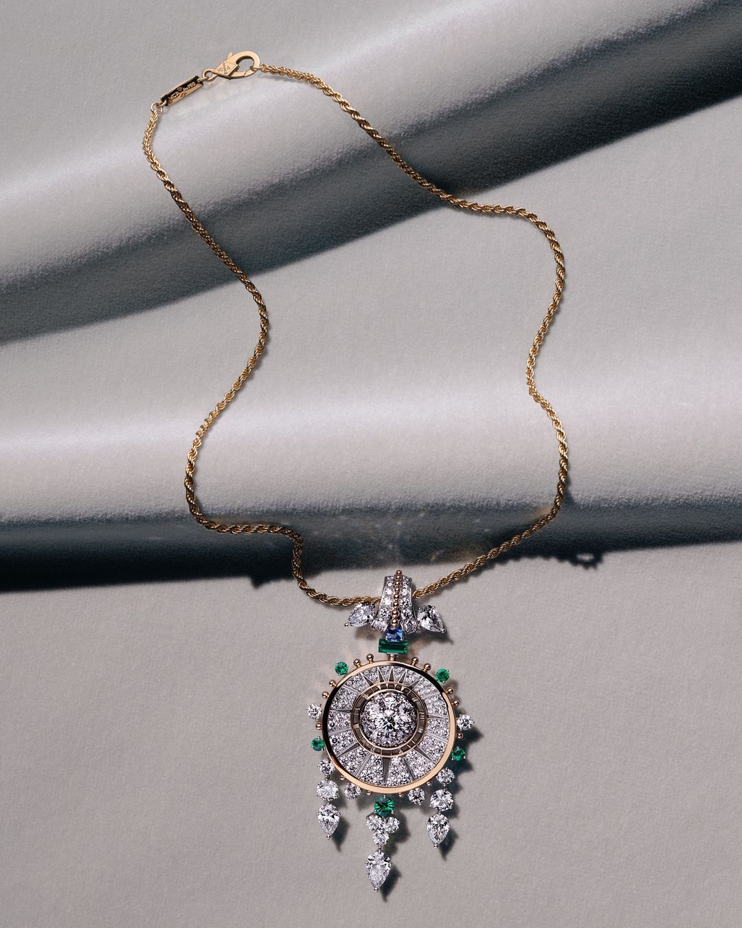 Gold chain necklace with a detailed diamond and gemstone pendant resting on a grey textured fabric background.
