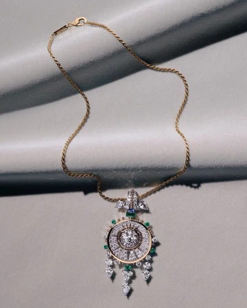 Gold chain necklace with a detailed diamond and gemstone pendant resting on a grey textured fabric background.