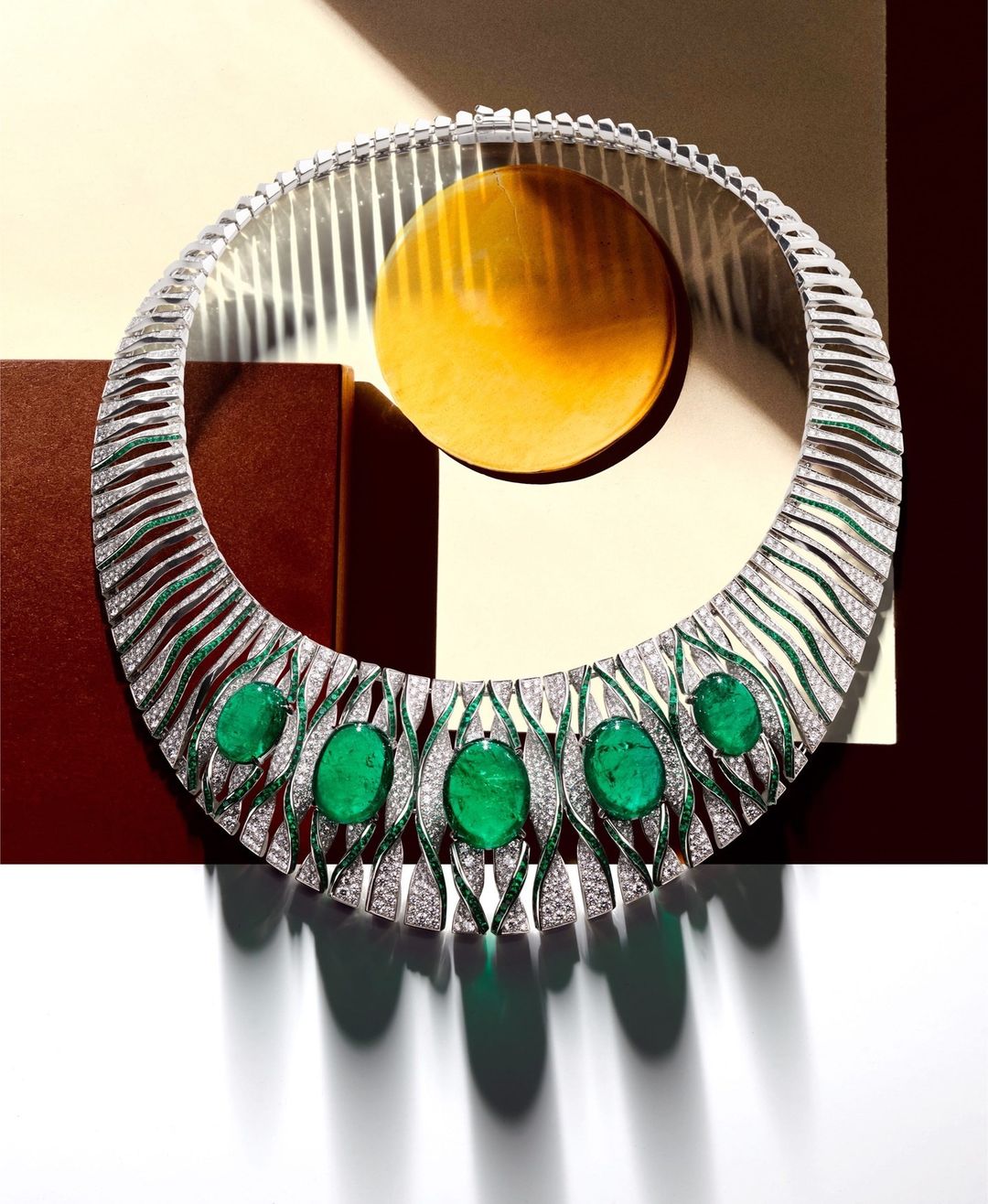 Elegant emerald and diamond collar necklace featuring green gemstones and white diamonds, displayed against a modern geometric background.