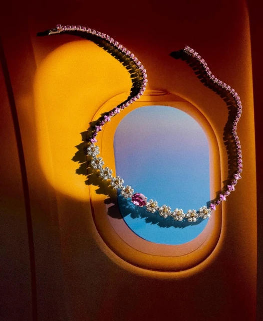 Elegant pink sapphire and diamond necklace displayed inside an airplane window with a gradient blue sky, bathed in warm, directed light.