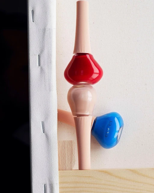 Still life of stacked nail polish bottles in red, beige, and blue against a white canvas background.