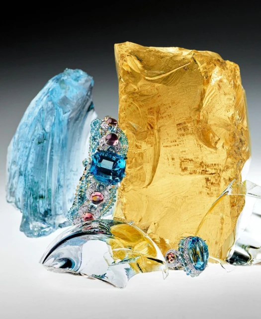 Still life shot of two bejeweled rings and a bracelet positioned against translucent gold and blue textured glass fragments in a studio setting.