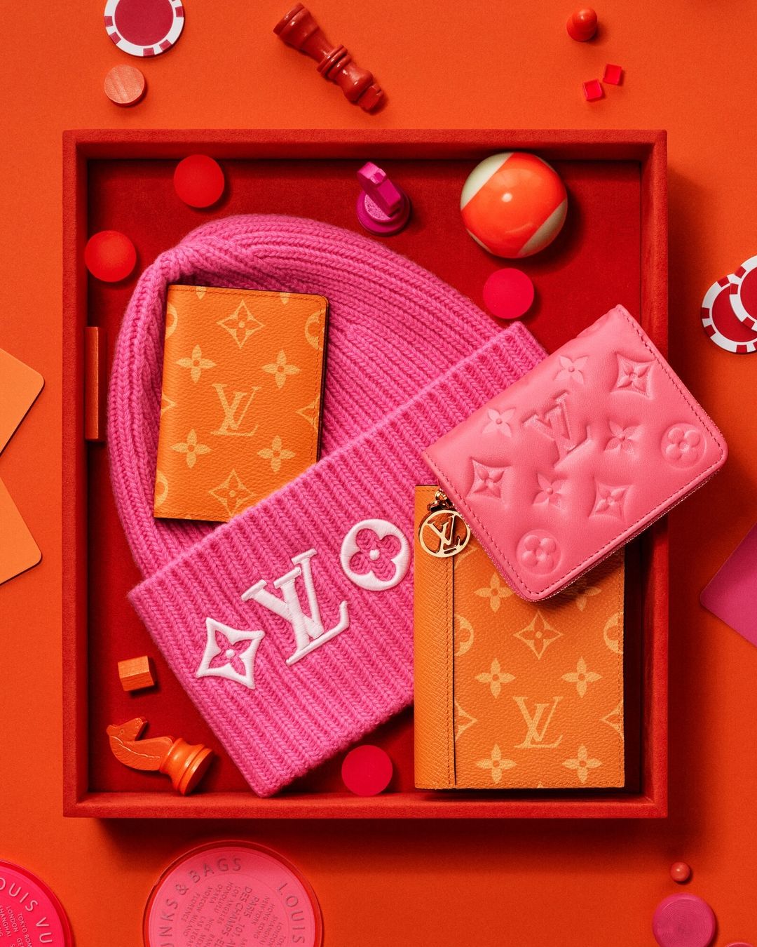 Elevated shot of Louis Vuitton wallets, cardholders, and a beanie in a vibrant orange and pink color palette on an orange background.