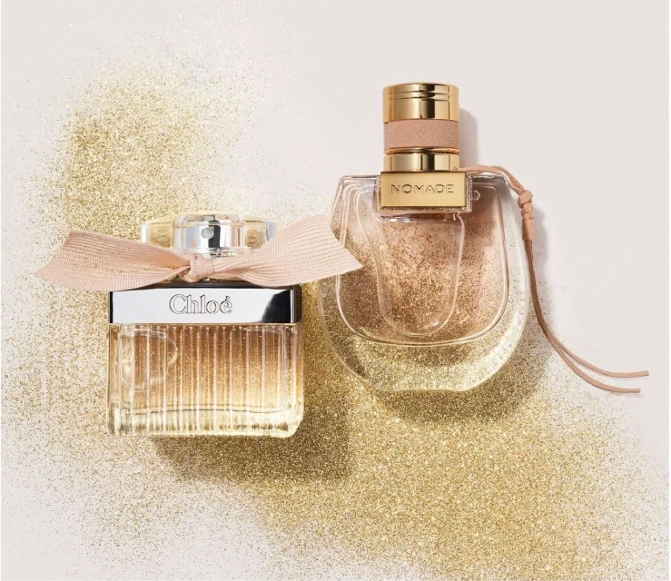 Two perfume bottles, Chloe and Nomade, sit on a sparkling gold surface. Chloe is rectangular with a bow, while Nomade is curved.