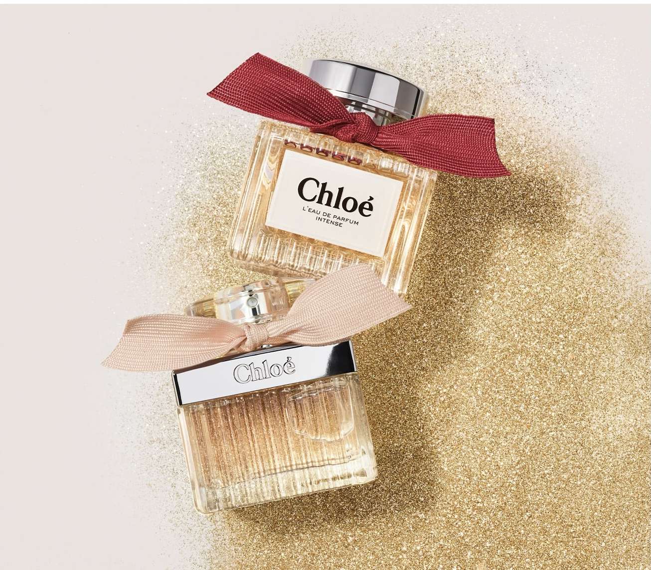 Two Chloe perfume bottles stacked on a gold glitter surface, one bottle with a dark red bow and the other with a light pink bow.