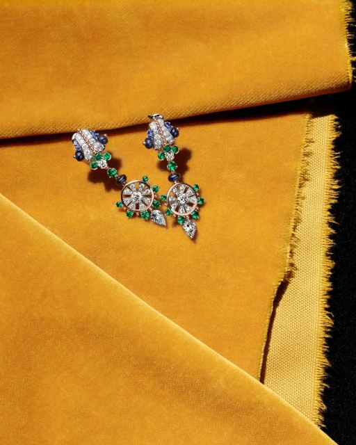 Elegant drop earrings with gemstones and diamonds displayed on a textured mustard fabric, jewellery still life, overhead shot.