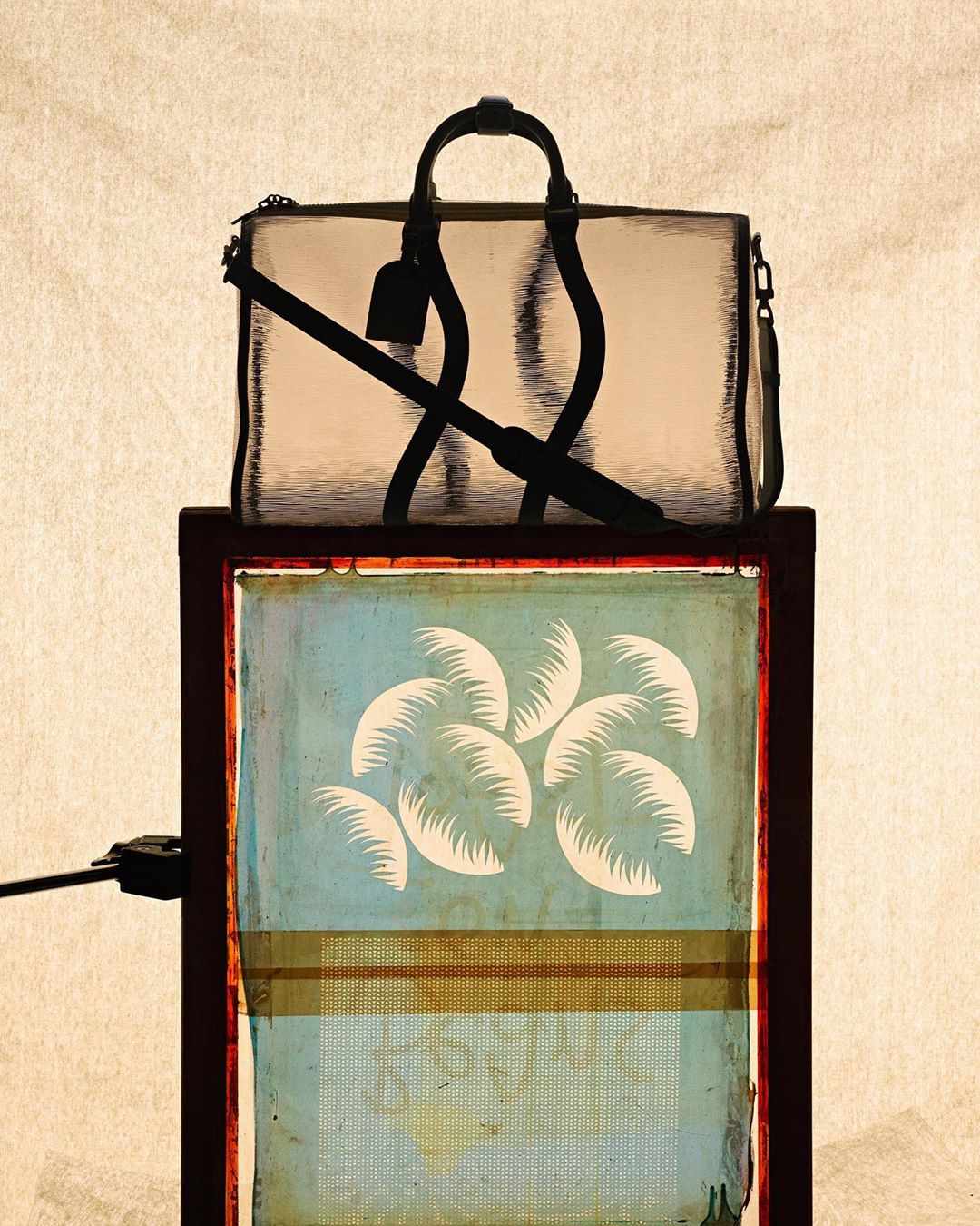 Studio shot of a handbag on top of an abstract framed artwork featuring palm leaf designs, set against a neutral textured background.