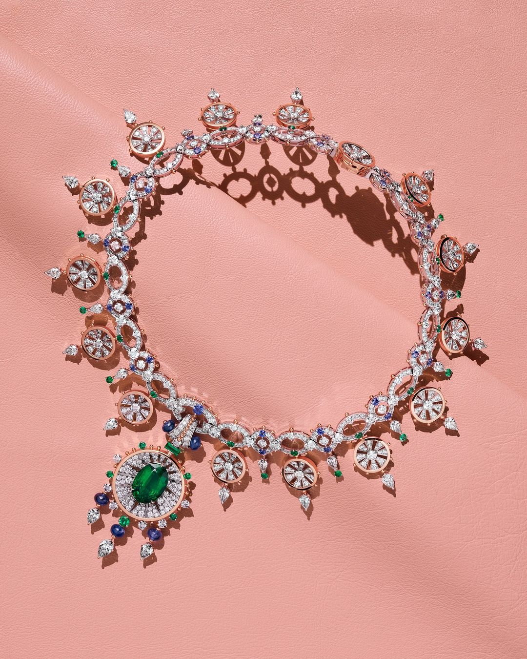 Elegant necklace with emerald, sapphire, and diamond gemstones against a pink leather backdrop. Jewelry design for fashion and beauty.