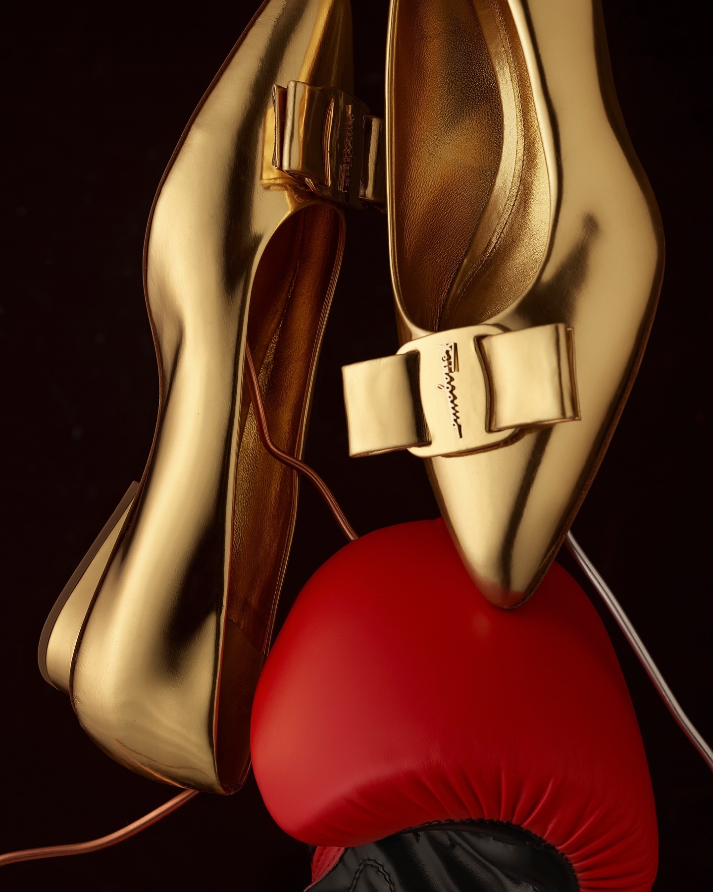 Elevated view of a pair of metallic gold high heels balanced on a red boxing glove against a dark backdrop. Fashion and sport.