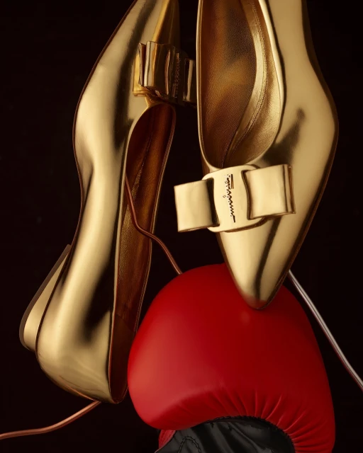 Elevated view of a pair of metallic gold high heels balanced on a red boxing glove against a dark backdrop. Fashion and sport.