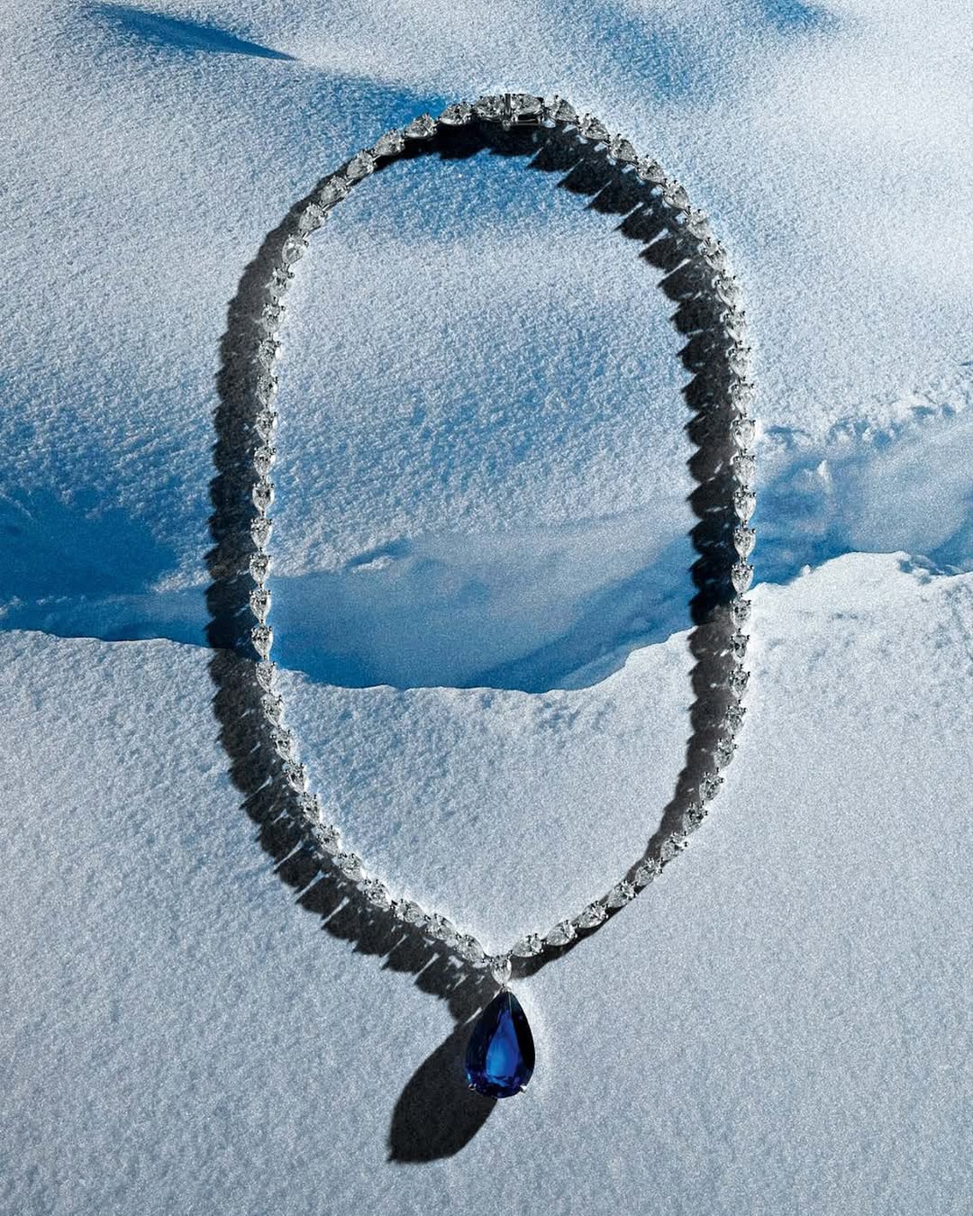 Elegant diamond necklace with teardrop sapphire pendant displayed on a snowy surface, highlighting luxury and natural contrast.