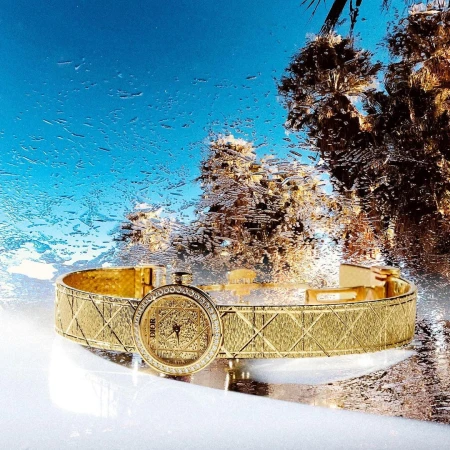 Luxurious gold cuff watch with diamond bezel resting on a reflective surface against a vibrant blue and palm tree backdrop with water droplets.