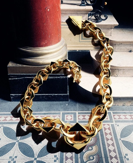 A bold gold chain necklace, with square and octagonal links, is showcased indoors next to a column and stairway, casting shadows on a mosaic tile floor.