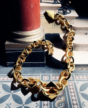 A bold gold chain necklace, with square and octagonal links, is showcased indoors next to a column and stairway, casting shadows on a mosaic tile floor.