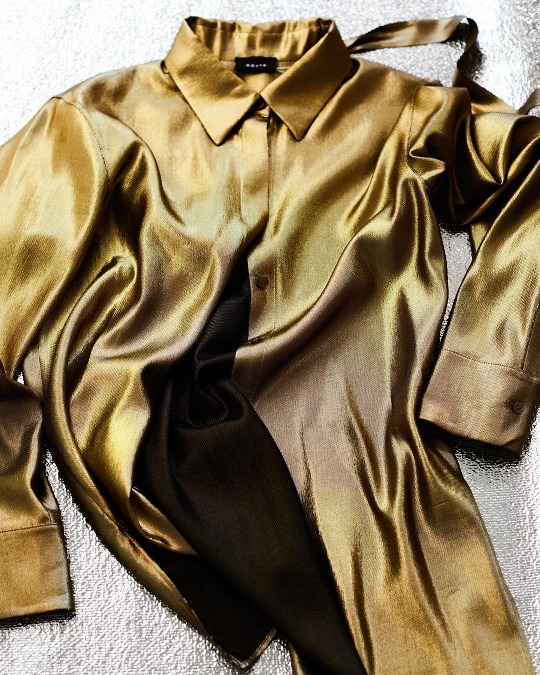 Satin gold-colored collared long-sleeve blouse with ties at the back of the neck, laid flat on a gray textured surface.