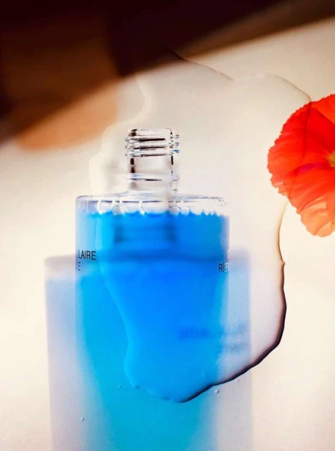 Close-up studio shot of a clear bottle with blue liquid, alongside a red flower and water droplets, on a cream-colored surface.