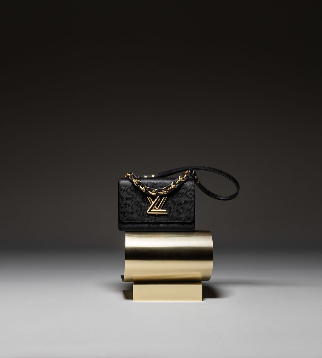 Luxury black leather Louis Vuitton purse with gold chain strap and gold hardware placed on a modern gold pedestal indoors.
