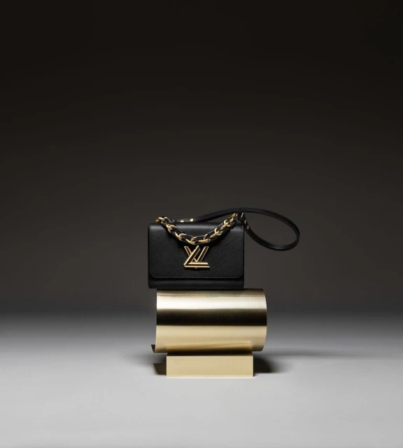 Luxury black leather Louis Vuitton purse with gold chain strap and gold hardware placed on a modern gold pedestal indoors.