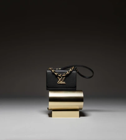 Luxury black leather Louis Vuitton purse with gold chain strap and gold hardware placed on a modern gold pedestal indoors.