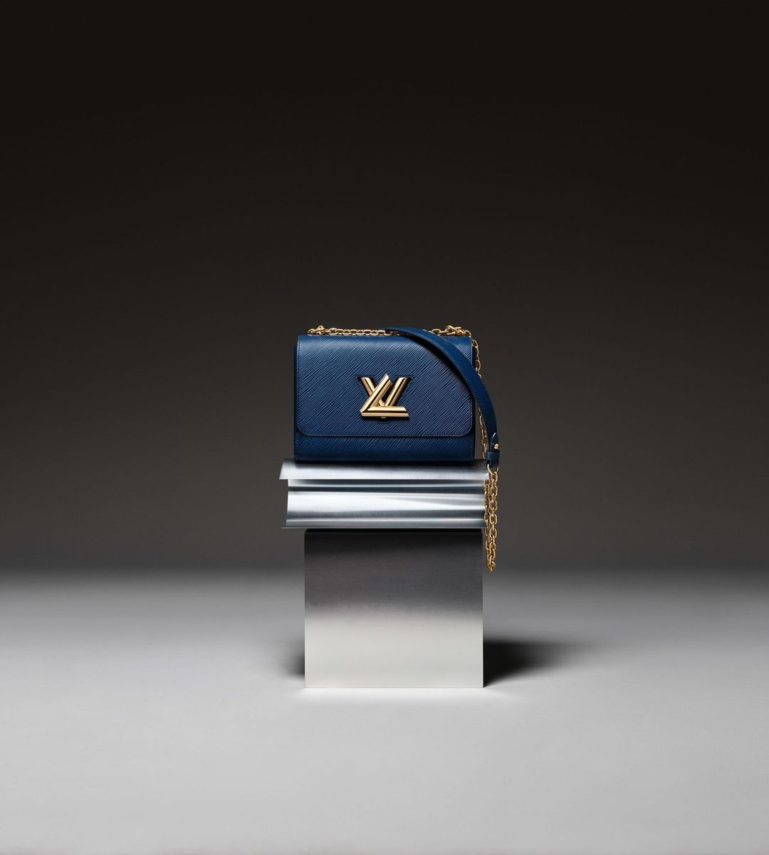 Luxury blue leather Louis Vuitton handbag with gold hardware and chain strap on a metallic pedestal against a dark background.