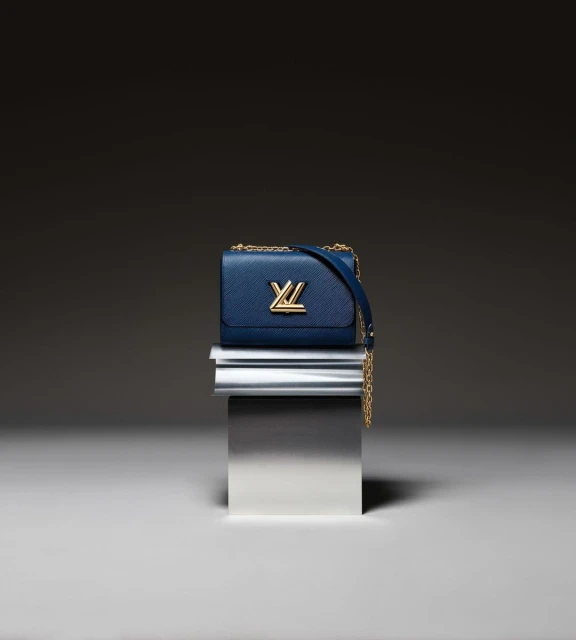 Luxury blue leather Louis Vuitton handbag with gold hardware and chain strap on a metallic pedestal against a dark background.