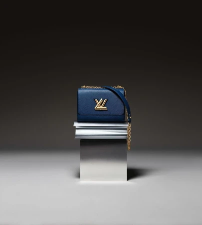 Luxury blue leather Louis Vuitton handbag with gold hardware and chain strap on a metallic pedestal against a dark background.