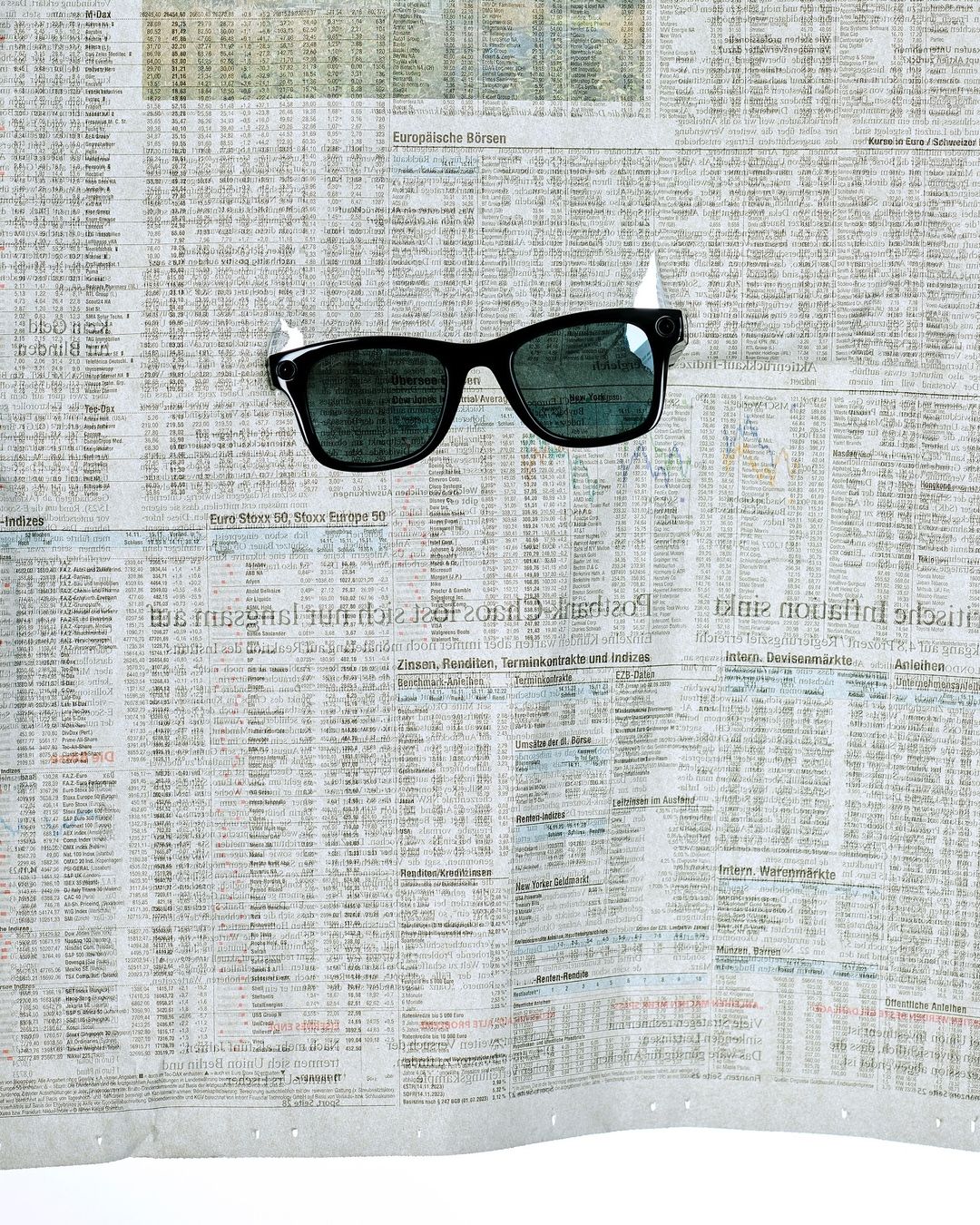 Black sunglasses taped to a crumpled newspaper sheet with financial data. The glasses are centered with tape visible at the top.